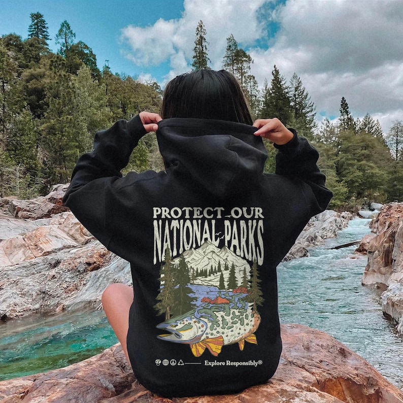 May include: A black hooded sweatshirt featuring a graphic on the back with the text "PROTECT OUR NATIONAL PARKS" above an illustration of a mountain range, pine trees, a river, and a large fish, with "Explore Responsibly" at the bottom.