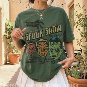 May include: A forest green short-sleeve t-shirt featuring a graphic print with the text Midnight Spook Show, Ft. Real Monsters, On Stage, The Creepiest Horror Show You've Ever Seen, and Friday Oct. 13, along with illustrations of three monster faces.
