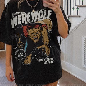 May include: A black short-sleeve t-shirt featuring a graphic of a werewolf with red and brown fur. The text reads: ONE NIGHT ONLY FT. WEREWOLF CURSE OF THE MOON, SIGHTINGS REPORTED SINCE 1200S NOW SEE UPCLOSE, and SOME LEGENDS ARE REAL.