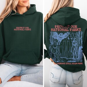 May include: A dark green hooded sweatshirt featuring the text "PROTECT OUR NATIONAL PARKS" in red on the front. The back displays a blue graphic of a forest scene with a large creature, an owl, and ghost-like figures with the text "LEGENDS STILL ROAM FREE."