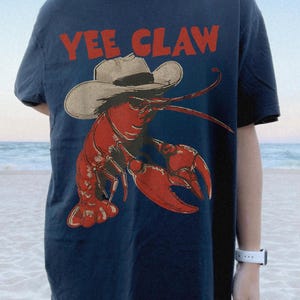 Op de afbeelding: Marineblauw T-shirt met een rode kreeft met een cowboyhoed. De tekst "YEE CLAW" is in het rood boven de kreeftafbeelding gedrukt. Het shirt heeft een casual stijl.