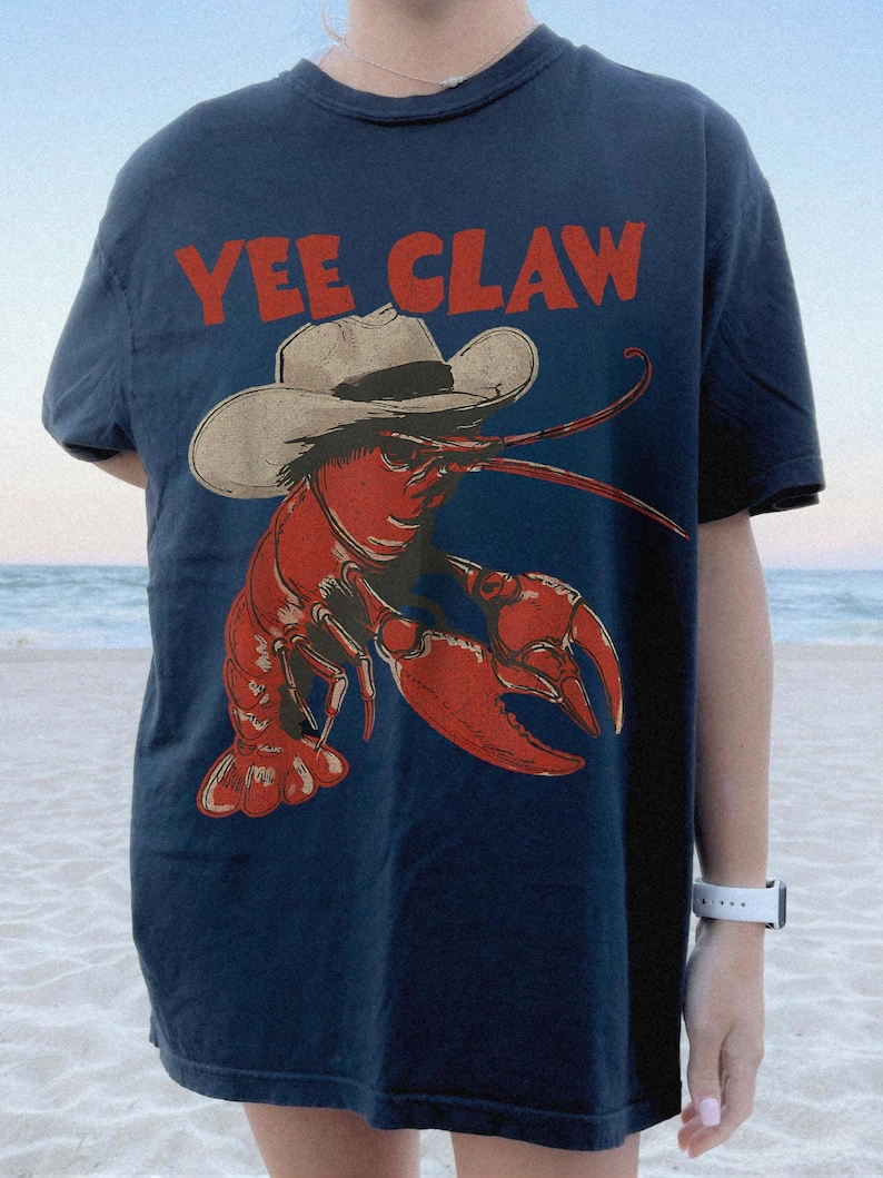 Op de afbeelding: Marineblauw T-shirt met een rode kreeft met een cowboyhoed. De tekst "YEE CLAW" is in het rood boven de kreeftafbeelding gedrukt. Het shirt heeft een casual stijl.
