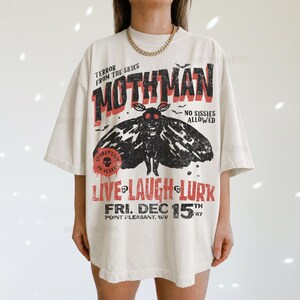 May include: An off-white short-sleeve t-shirt featuring a black moth graphic and red text that reads TERROR FROM THE SKIES MOTHMAN NO SISSIES ALLOWED LIVE LAUGH LURK FRI. DEC 15TH POINT PLEASANT, WV. The design has a distressed, vintage-inspired aesthetic.