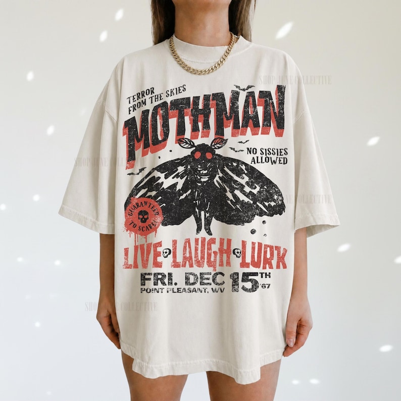 May include: An off-white short-sleeve t-shirt featuring a black moth graphic and red text that reads TERROR FROM THE SKIES MOTHMAN NO SISSIES ALLOWED LIVE LAUGH LURK FRI. DEC 15TH POINT PLEASANT, WV. The design has a distressed, vintage-inspired aesthetic.