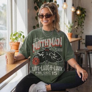 May include: An olive green t-shirt featuring a graphic of a moth with red eyes. The text reads: TERROR FROM THE SKIES, MOTHMAN, NO SISSIES ALLOWED, LIVE LAUGH LURK, FRI. DEC 15TH 67, POINT PLEASANT, WV. A red circular stamp says GUARANTEED TO SCARE.