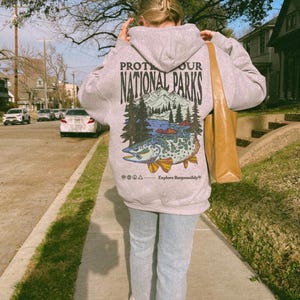 May include: A light gray hooded sweatshirt featuring a graphic on the back with the text "PROTECT OUR NATIONAL PARKS" above an illustration of mountains, pine trees, and a large fish, with the text "Explore Responsibly" at the bottom.