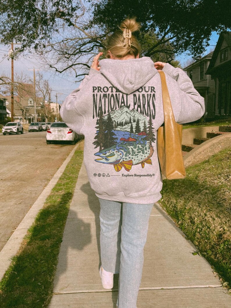 May include: A light gray hooded sweatshirt featuring a graphic on the back with the text "PROTECT OUR NATIONAL PARKS" above an illustration of mountains, pine trees, and a large fish, with the text "Explore Responsibly" at the bottom.
