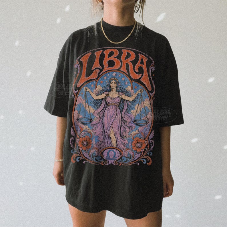 May include: A black short-sleeve t-shirt featuring a large graphic print on the front. The design displays the word LIBRA in orange letters above an illustration of a figure holding scales, surrounded by blue clouds, orange flowers, and swirls.