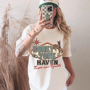 Pode incluir: Camiseta creme com a frase "Honky Tonk Haven Tunes and Tequila". O design apresenta um cowboy em um cavalo selvagem e uma estrela vermelha. Um bon&eacute; de beisebol bege e verde tamb&eacute;m &eacute; vis&iacute;vel.