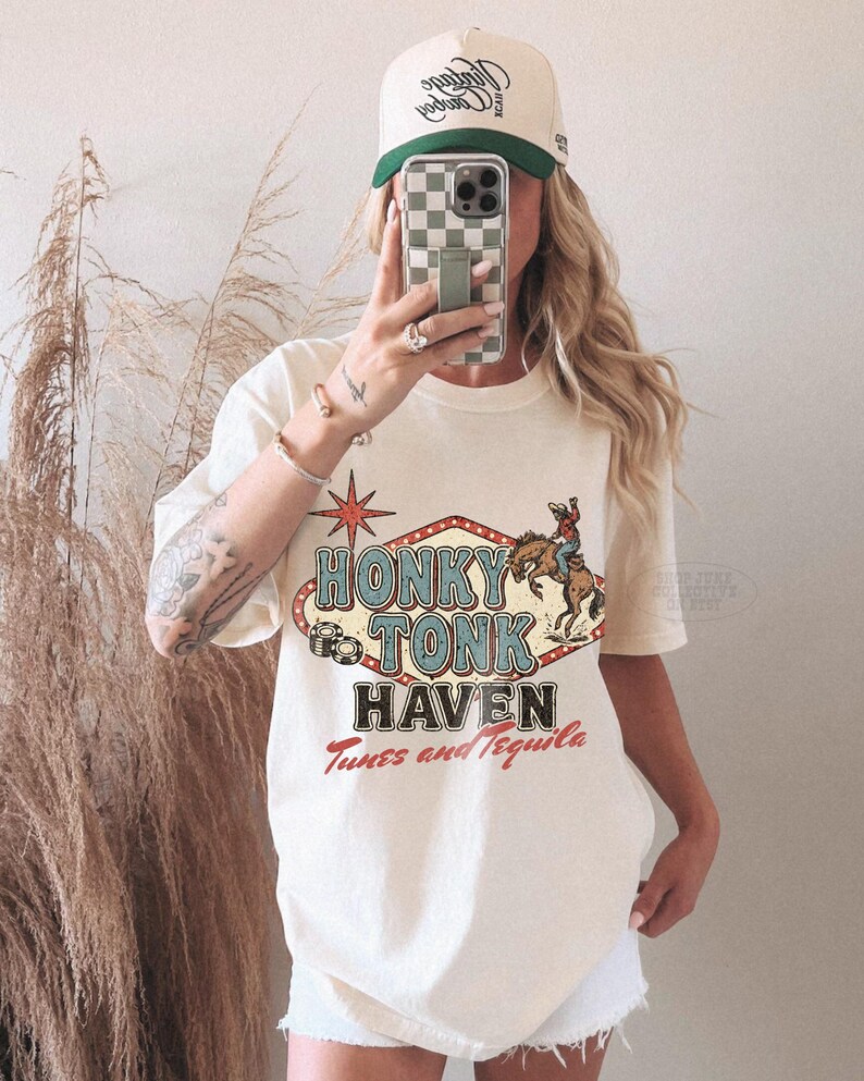 Pode incluir: Camiseta creme com a frase "Honky Tonk Haven Tunes and Tequila". O design apresenta um cowboy em um cavalo selvagem e uma estrela vermelha. Um bon&eacute; de beisebol bege e verde tamb&eacute;m &eacute; vis&iacute;vel.