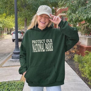 May include: A dark green hooded sweatshirt featuring the text PROTECT OUR NATIONAL PARKS in a cream-colored serif font on the chest. The garment includes a front kangaroo pocket and long sleeves, paired with a light beige baseball cap.