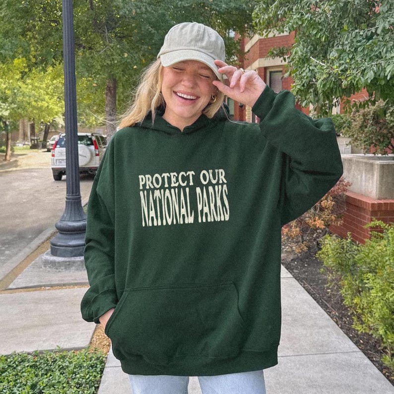 May include: A dark green hooded sweatshirt featuring the text PROTECT OUR NATIONAL PARKS in a cream-colored serif font on the chest. The garment includes a front kangaroo pocket and long sleeves, paired with a light beige baseball cap.