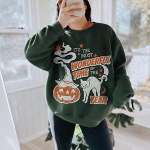 Peut inclure: Un sweat-shirt &agrave; col rond vert fonc&eacute; avec une impression graphique portant le texte "IT'S THE MOST WONDERFUL TIME OF THE YEAR" en orange et blanc. Le motif comprend un fant&ocirc;me blanc, une citrouille avec un chapeau, un chat noir et des &eacute;toiles.