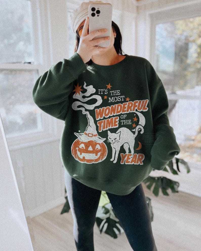 Peut inclure: Un sweat-shirt &agrave; col rond vert fonc&eacute; avec une impression graphique portant le texte "IT'S THE MOST WONDERFUL TIME OF THE YEAR" en orange et blanc. Le motif comprend un fant&ocirc;me blanc, une citrouille avec un chapeau, un chat noir et des &eacute;toiles.