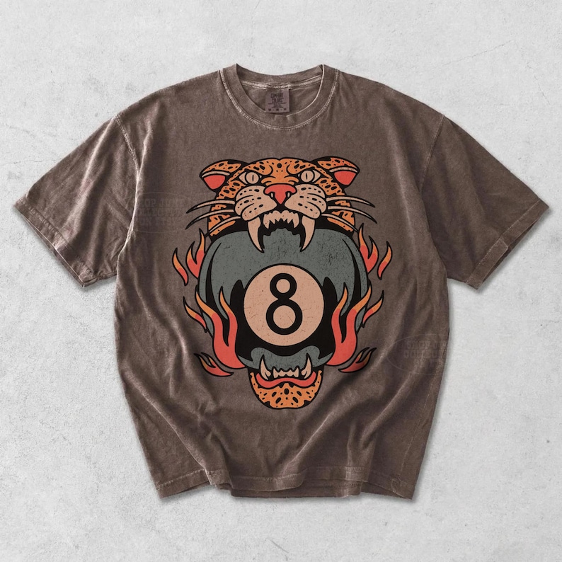 May include: Brown t-shirt featuring a graphic of a tiger's head biting an eight ball, surrounded by flames. The design is in shades of orange, red, and blue. The shirt has a vintage, worn look.