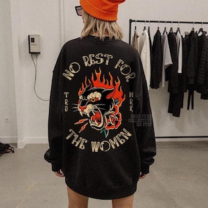 May include: Black sweatshirt with a graphic of a panther, rose, and lightning bolt, surrounded by flames. The text "NO REST FOR THE WOMEN" is arched above the graphic. The sweatshirt is worn with an orange beanie.