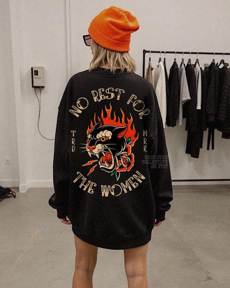 May include: Black sweatshirt with a graphic of a panther, rose, and lightning bolt, surrounded by flames. The text "NO REST FOR THE WOMEN" is arched above the graphic. The sweatshirt is worn with an orange beanie.