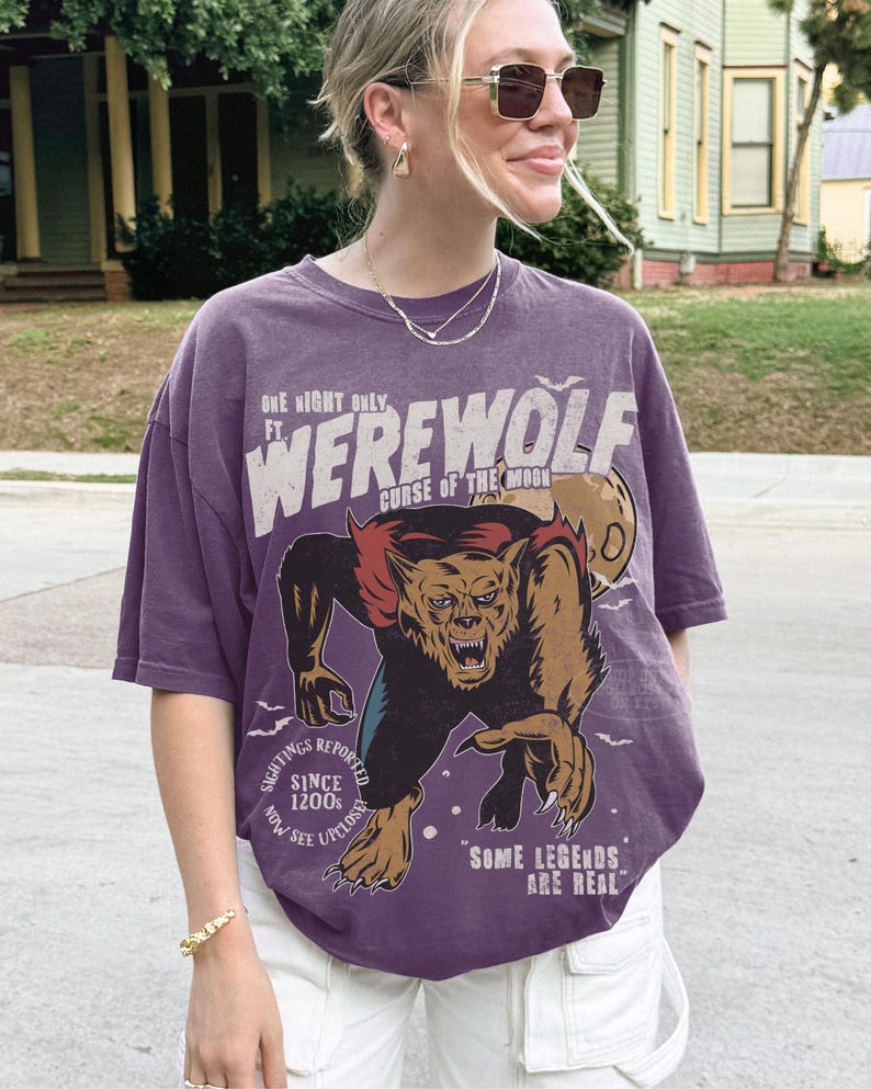 May include: A purple short-sleeve t-shirt featuring a graphic of a werewolf with the text "ONE NIGHT ONLY FT. WEREWOLF CURSE OF THE MOON", "SIGHTINGS REPORTED SINCE 1200s NOW SEE UPCLOSE", and "SOME LEGENDS ARE REAL" printed in white and tan.