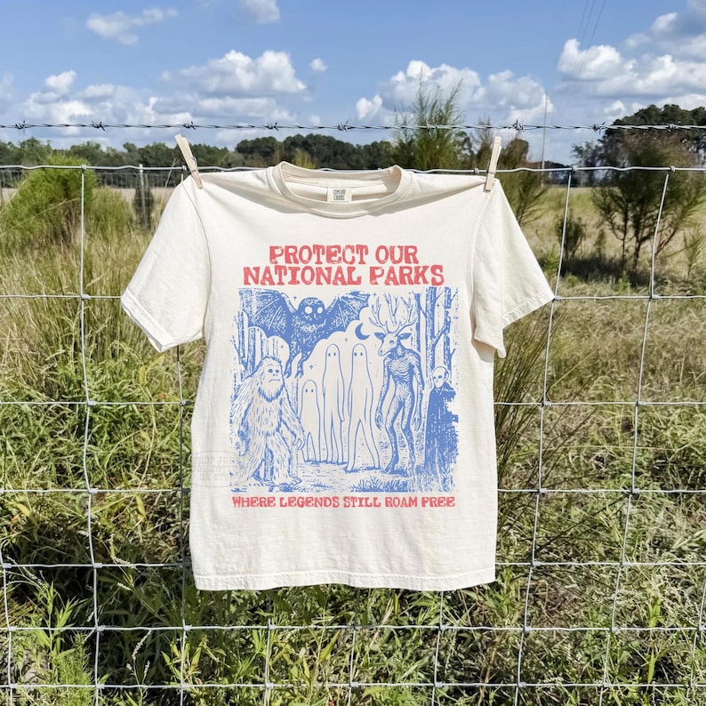 Peut inclure: Un t-shirt &agrave; manches courtes couleur cr&egrave;me avec une impression graphique bleue repr&eacute;sentant une chauve-souris, un sasquatch, des fant&ocirc;mes, une cr&eacute;ature ressemblant &agrave; un cerf et une for&ecirc;t. Le texte indique PROTECT OUR NATIONAL PARKS et WHERE LEGENDS STILL ROAM FREE en rouge.
