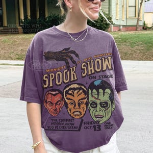 May include: A purple short-sleeve t-shirt featuring a graphic print with the text Midnight Spook Show, Ft. Real Monsters, On Stage, The Creepiest Horror Show You've Ever Seen, Friday Oct. 13, and One Night Only, alongside illustrations of three monster faces.