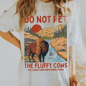 May include: A white t-shirt featuring a vintage-style graphic of a bison in Yellowstone National Park. The graphic reads "Do Not Pet The Fluffy Cows Yellowstone National Park".