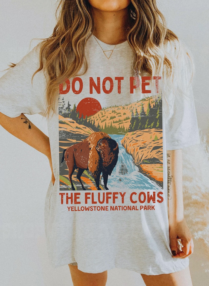 May include: A white t-shirt featuring a vintage-style graphic of a bison in Yellowstone National Park. The graphic reads "Do Not Pet The Fluffy Cows Yellowstone National Park".