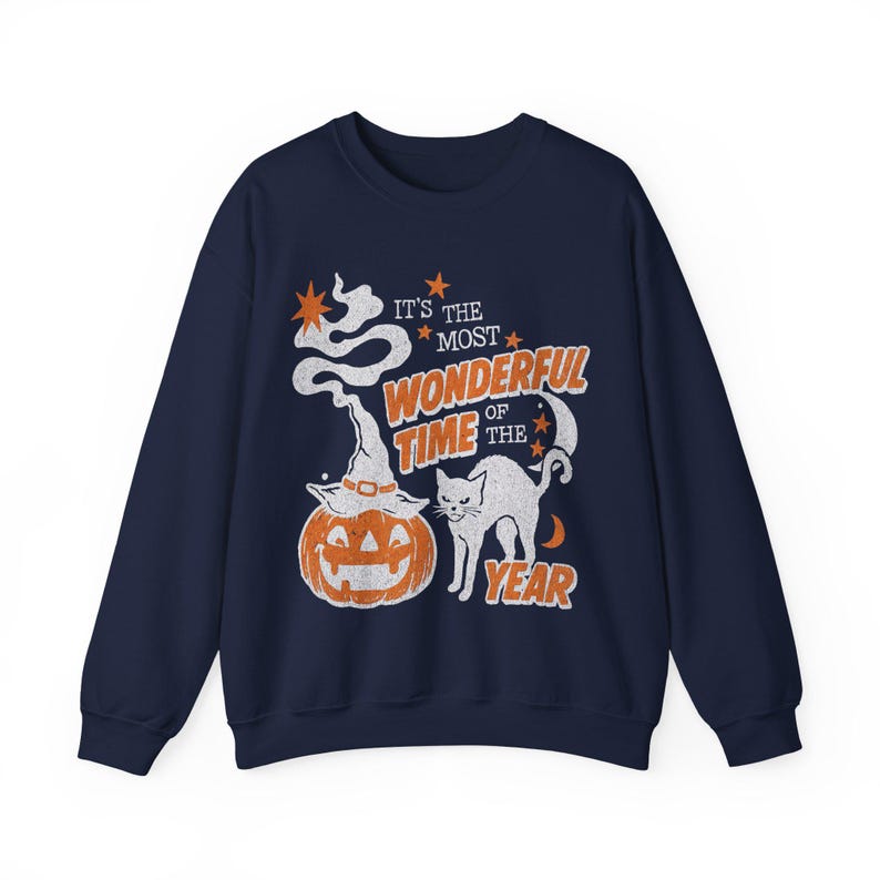 Peut inclure: Un sweat-shirt bleu marine &agrave; col rond avec une impression graphique vieillie repr&eacute;sentant une citrouille d'Halloween portant un chapeau de sorci&egrave;re, un chat noir, des &eacute;toiles et un croissant de lune. Le texte indique IT'S THE MOST WONDERFUL TIME OF THE YEAR.