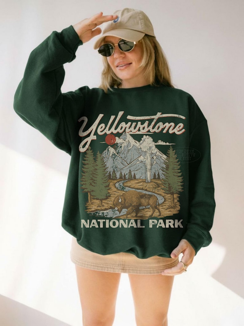 May include: A dark green crewneck sweatshirt featuring a graphic print of Yellowstone National Park. The design includes a bison, a winding river, pine trees, mountains, and a red sun, with the text Yellowstone National Park in a vintage font.