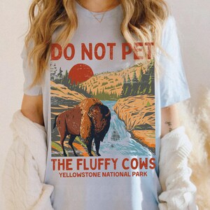 May include: A light blue t-shirt featuring a vintage-style graphic of a bison in Yellowstone National Park. The graphic reads "Do Not Pet The Fluffy Cows".