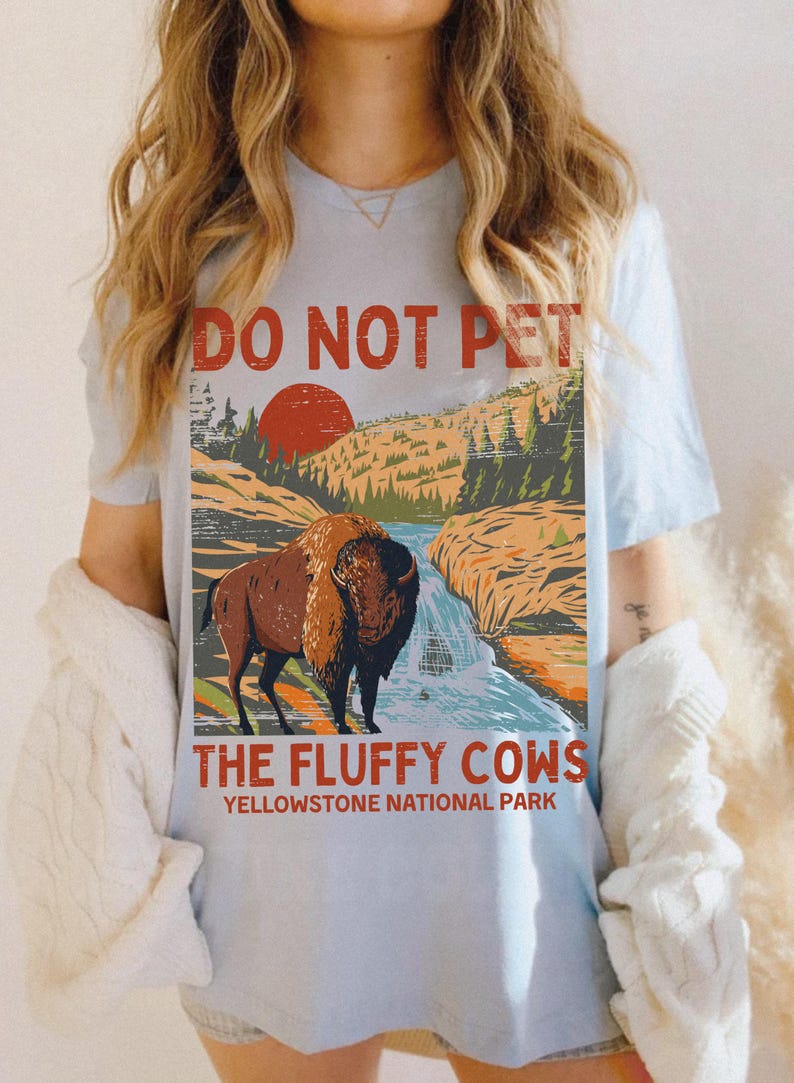 May include: A light blue t-shirt featuring a vintage-style graphic of a bison in Yellowstone National Park. The graphic reads "Do Not Pet The Fluffy Cows".