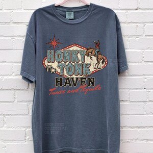Pode incluir: Uma camiseta azul com um design gr&aacute;fico retr&ocirc; "Honky Tonk Haven". O design inclui um cowboy em um cavalo selvagem e uma estrela vermelha. A camiseta tamb&eacute;m tem o texto "Tunes and Tequila".