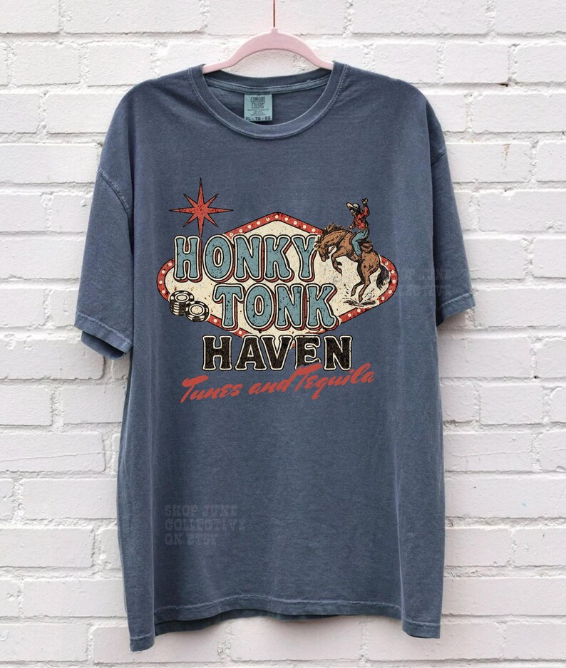 Pode incluir: Uma camiseta azul com um design gr&aacute;fico retr&ocirc; "Honky Tonk Haven". O design inclui um cowboy em um cavalo selvagem e uma estrela vermelha. A camiseta tamb&eacute;m tem o texto "Tunes and Tequila".