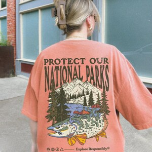 May include: A coral-colored t-shirt with a vintage-style graphic of a trout, mountains, and trees. The text "Protect Our National Parks" is printed in brown on the shirt. The text "Explore Responsibly" is printed in brown at the bottom of the graphic.