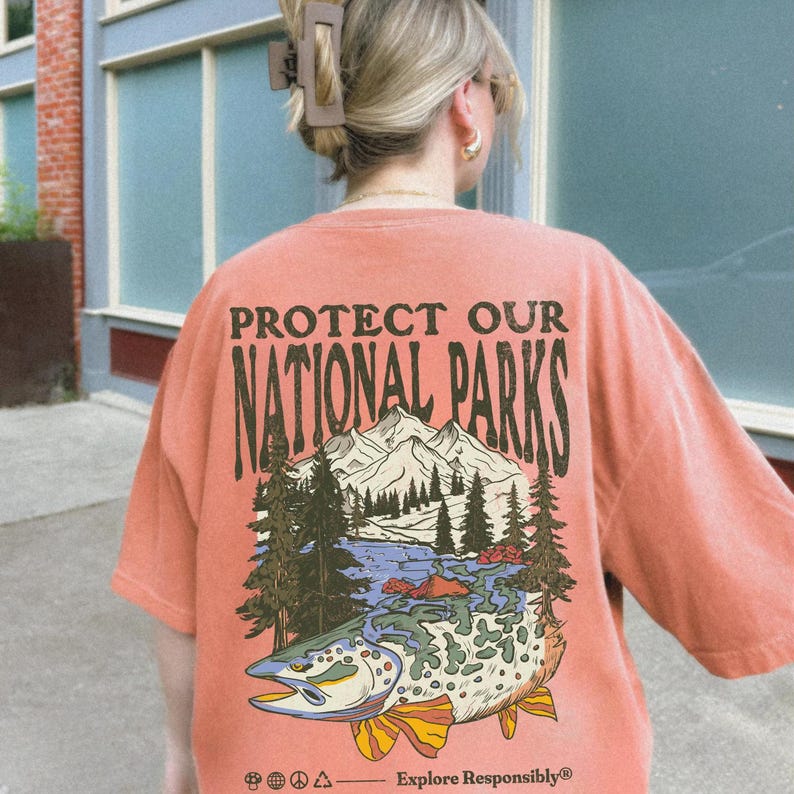 May include: A coral-colored t-shirt with a vintage-style graphic of a trout, mountains, and trees. The text "Protect Our National Parks" is printed in brown on the shirt. The text "Explore Responsibly" is printed in brown at the bottom of the graphic.