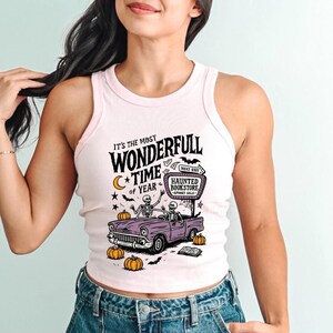 May include: A light pink tank top with a graphic design. The design features a purple car with skeleton drivers, pumpkins, and the text "It's the Most Wonderfull Time of Year" and "Haunted Bookstore".