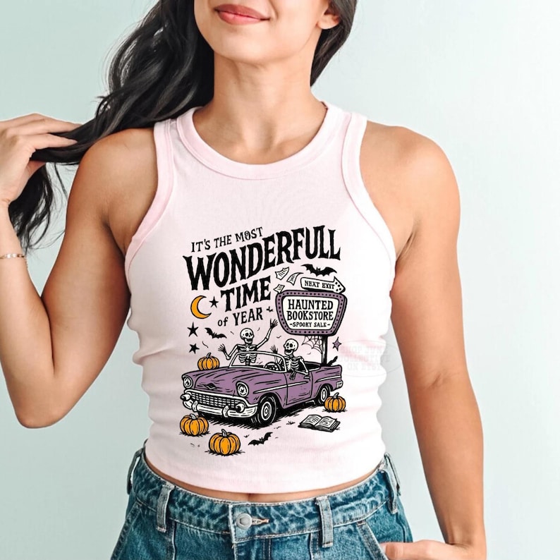 May include: A light pink tank top with a graphic design. The design features a purple car with skeleton drivers, pumpkins, and the text "It's the Most Wonderfull Time of Year" and "Haunted Bookstore".