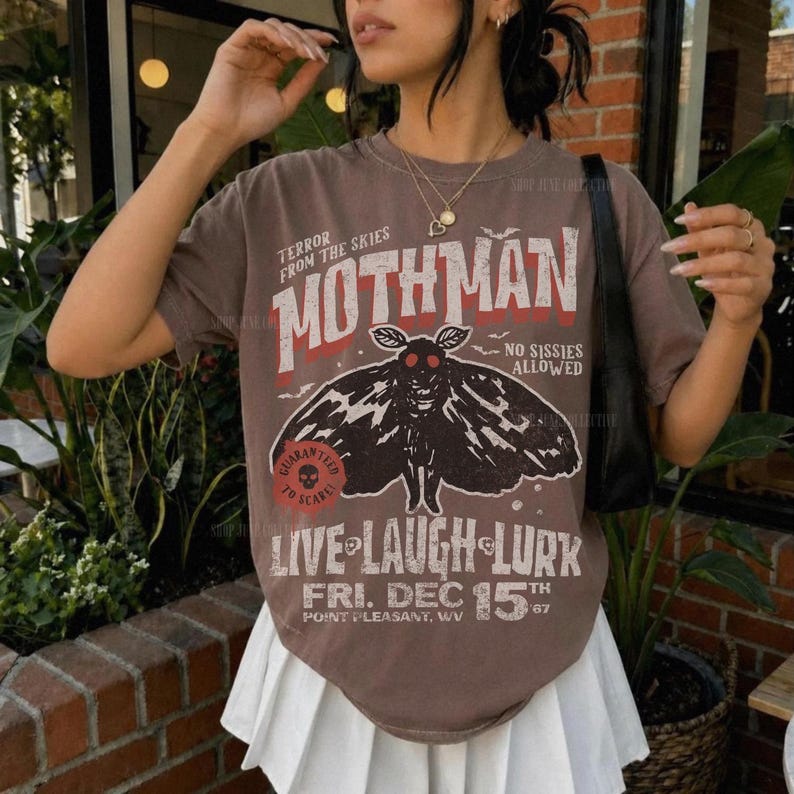 May include: A brown short-sleeve t-shirt featuring a graphic of a moth with red eyes. The text reads: TERROR FROM THE SKIES MOTHMAN NO SISSIES ALLOWED LIVE LAUGH LURK FRI. DEC 15TH POINT PLEASANT, WV. The shirt is paired with a white pleated skirt.