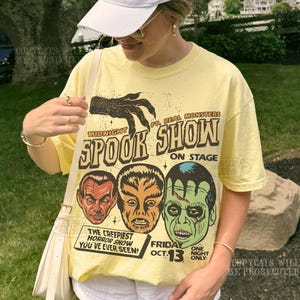 May include: A pale yellow short-sleeve t-shirt featuring a graphic print of three monster faces and a clawed hand. The text reads: Midnight Spook Show ft. Real Monsters on stage, The Creepiest Horror Show You've Ever Seen! Friday Oct. 13.