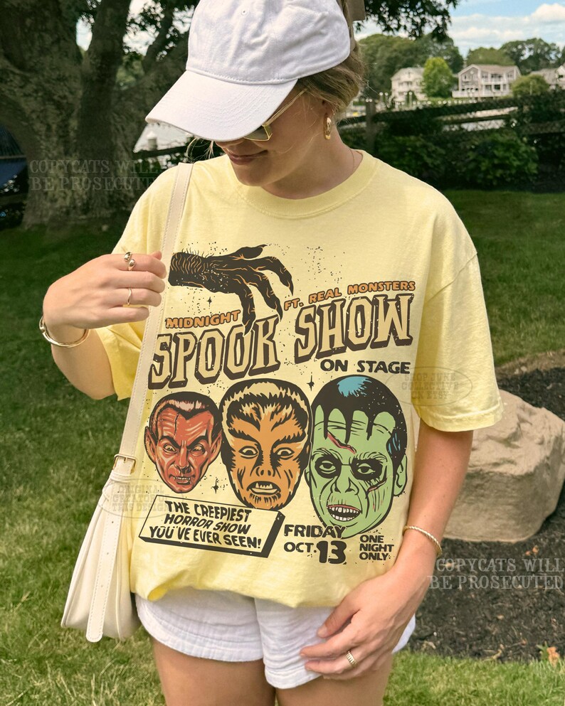May include: A pale yellow short-sleeve t-shirt featuring a graphic print of three monster faces and a clawed hand. The text reads: Midnight Spook Show ft. Real Monsters on stage, The Creepiest Horror Show You've Ever Seen! Friday Oct. 13.