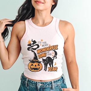 May include: A light pink tank top with a Halloween-themed graphic. The graphic includes a jack-o'-lantern wearing a witch hat, a black cat, and the text "It's the most wonderful time of the year."