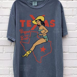 Op de afbeelding: Blauw-grijs T-shirt met een retro-afbeelding. Het toont een vrouw in badpak en cowboylaarzen, tegen een rode omtrek van Texas. De tekst luidt "TEXAS Biggest and Best".