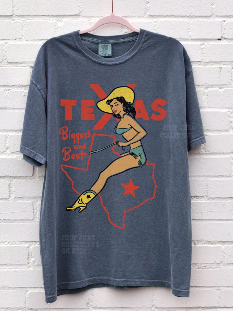 Op de afbeelding: Blauw-grijs T-shirt met een retro-afbeelding. Het toont een vrouw in badpak en cowboylaarzen, tegen een rode omtrek van Texas. De tekst luidt "TEXAS Biggest and Best".