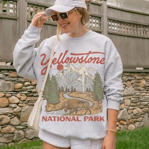 May include: A light gray crewneck sweatshirt featuring a graphic print of Yellowstone National Park with a bison, pine trees, mountains, and a geyser. The text Yellowstone National Park is printed in red. A white baseball cap and sunglasses.