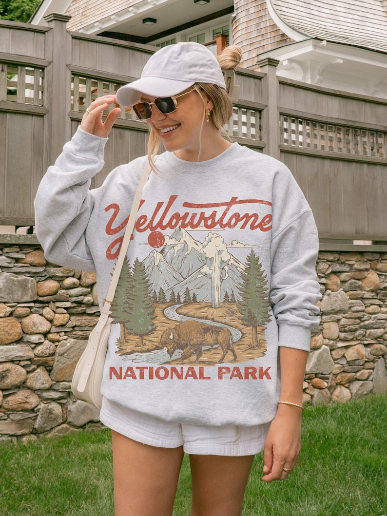 May include: A light gray crewneck sweatshirt featuring a graphic print of Yellowstone National Park with a bison, pine trees, mountains, and a geyser. The text Yellowstone National Park is printed in red. A white baseball cap and sunglasses.