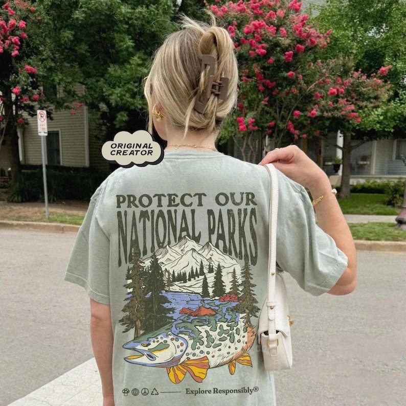 May include: A light green t-shirt with a graphic print of a fish and a mountain scene. The text "Protect Our National Parks" is printed on the shirt. The text "Explore Responsibly" is printed below the graphic.