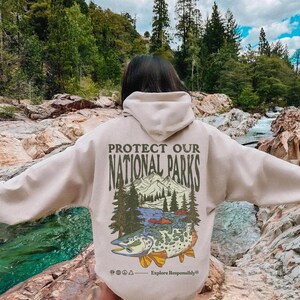 May include: A beige hooded sweatshirt featuring a back graphic with the text PROTECT OUR NATIONAL PARKS and Explore Responsibly. The design includes a mountain range, pine trees, and a large fish with a detailed pattern, plus small icons.