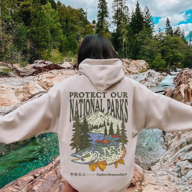 May include: A beige hooded sweatshirt featuring a back graphic with the text PROTECT OUR NATIONAL PARKS and Explore Responsibly. The design includes a mountain range, pine trees, and a large fish with a detailed pattern, plus small icons.
