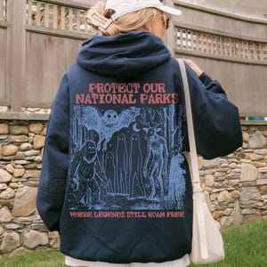 May include: A navy blue hooded sweatshirt featuring a graphic print on the back with the text "PROTECT OUR NATIONAL PARKS" and "WHERE LEGENDS STILL ROAM FREE" in red, alongside blue illustrations of an owl, a sasquatch, ghosts, and a deer-like creature.