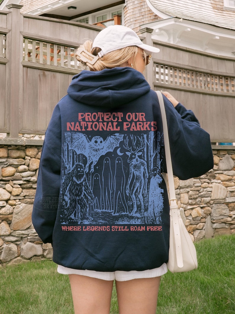 May include: A navy blue hooded sweatshirt featuring a graphic print on the back with the text "PROTECT OUR NATIONAL PARKS" and "WHERE LEGENDS STILL ROAM FREE" in red, alongside blue illustrations of an owl, a sasquatch, ghosts, and a deer-like creature.