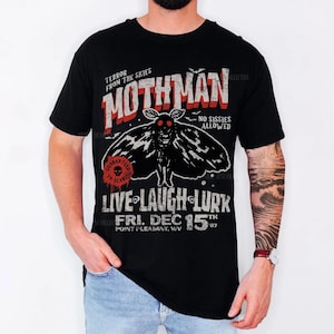 May include: A black t-shirt featuring a graphic of a moth with red eyes. The text reads: TERROR FROM THE SKIES, MOTHMAN, NO SISSIES ALLOWED, LIVE LAUGH LURK, FRI. DEC 15TH 67, POINT PLEASANT, WV. A red circular stamp says GUARANTEED TO SCARE.